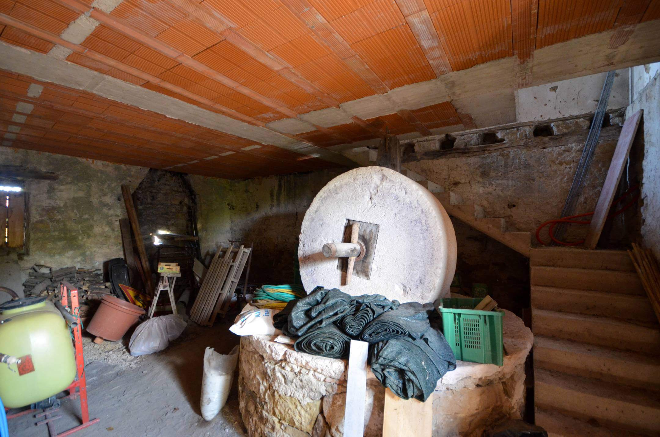 Prašna kletna delavnica z velikim okroglim brusilnim kolesom, opečnatim stropom ter raztresenim orodjem in cevmi.